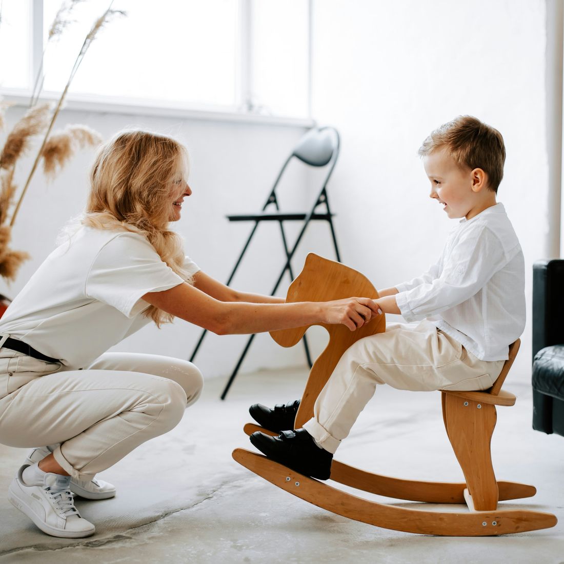 A woman playing with a smiling young boy on a wooden rocking horse in a bright room